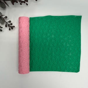 May include: A pink rolling pin and a green mat, both featuring a repeating leaf pattern. The rolling pin is cylindrical and rolled up, while the mat is rectangular and unrolled. The leaf pattern is embossed on both items.