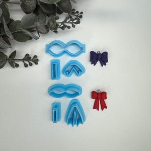 May include: A set of blue plastic cookie cutters in the shapes of glasses, bows, and a pair of wings. There are also two red and purple bows.