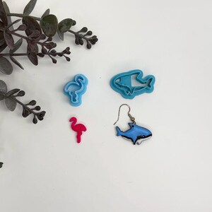 May include: A light blue flamingo cookie cutter, a light blue shark cookie cutter, a small pink flamingo charm, and a blue and white shark earring with a silver-toned metal hook. These items are arranged together on a plain white surface.