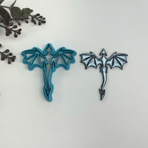 May include: Two dragon cookie cutters, one blue and one silver. The blue cutter is a detailed silhouette of a dragon with wings spread, while the silver cutter is a more stylized version with a simpler design.