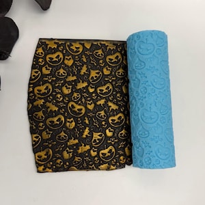 May include: A black and gold Halloween-themed clay sheet with a repeating pattern of jack-o'-lanterns, bats, and other spooky shapes. A blue roller with a matching pumpkin pattern is next to the sheet.