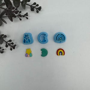 May include: A collection of six colorful polymer clay cutters in the shapes of a star, crescent moon, and rainbow. The cutters are in shades of blue, green, yellow, and rainbow colors. The cutters are for crafting and creating decorative elements.