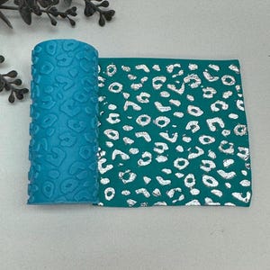 May include: A rolled turquoise material with a raised leopard print pattern. Next to it is a flat piece of teal material with a silver leopard print design. The items are displayed on a light gray surface.