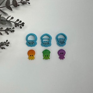 May include: A collection of blue plastic jellyfish-shaped cookie cutters and three colorful octopus-shaped cutters in orange, green, and purple. The cutters are arranged on a white surface.