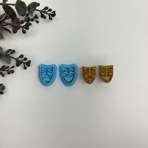 May include: Two pairs of theater mask-shaped earrings. The first pair is light blue, and the second pair is gold. The earrings are small and detailed, with a classic comedy and tragedy mask design. The background is white.