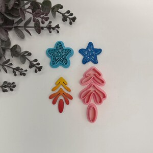May include: Four colorful polymer clay cutters in the shape of a starfish, a tree, and a leaf. The starfish cutters are blue, and the tree and leaf cutters are pink and yellow. The cutters are for crafting or baking.