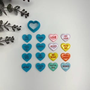 May include: A collection of colorful heart-shaped candies with various messages, including "LOVE," "HUGS," and "BE MINE." A blue heart-shaped cookie cutter is also present. The candies are in shades of pink, yellow, orange, and blue.