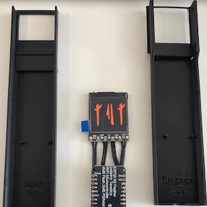 以下が含まれることがあります： 黒色の電子部品の俯瞰図。長方形のフレーム、テキスト付きの回路基板、オレンジ色の記号を表示する小さな画面が写っています。フレームには「CA2427」と「Engage C-51」の文字が記載されています。