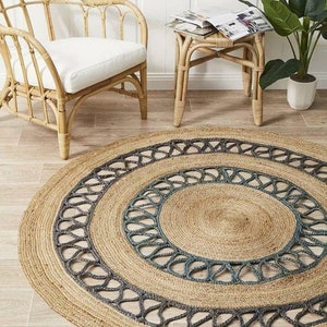 May include: A round jute rug with a natural tan color and a dark gray patterned border. The rug is placed on a light wood floor, with a rattan chair and side table in the background. The rug's diameter is approximately 60 inches.