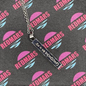 May include: A silver-colored pendant necklace with a rectangular charm. The charm has the text "Δv = ve ln(m0/mf)" in a black font. The background is a dark gray fabric with a repeating pattern of the word "REDMARS" in pink and blue.
