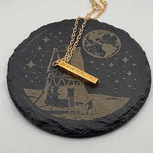 May include: A gold-colored bar pendant necklace with the formula "Δv = ve ln(mo / mf)" engraved on it. The necklace is displayed on a black, round coaster with a moon landing scene etched into it.