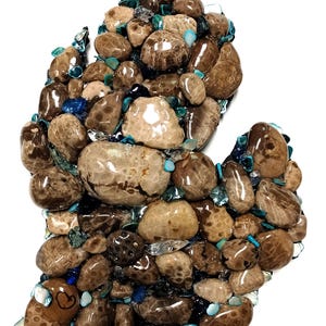 May include: A mosaic of brown, tan, and beige stones and pebbles arranged in the shape of a mitten. The stones are interspersed with blue and turquoise glass pieces. The artwork is set against a white background.