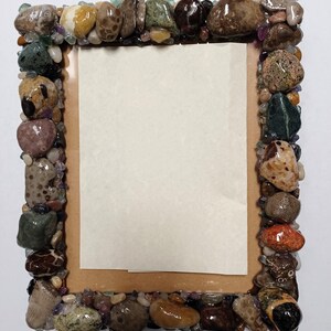 May include: A rectangular picture frame with a border made of various colorful stones and pebbles. The frame has a light brown backing and a blank, off-white paper insert. The stones are in shades of brown, green, and purple.