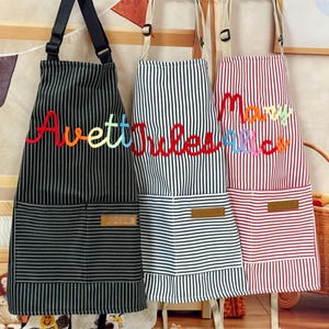 May include: Three striped aprons with adjustable neck straps and front pockets. The black apron features the embroidered name Avett, the white apron says Jules, and the red apron displays the embroidered names Mary Alice in colorful thread.
