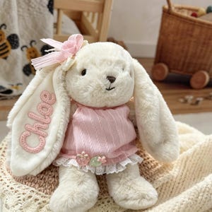 May include: A cream-colored plush rabbit toy with long ears, wearing a pink ribbed top with a white lace trim and two small strawberry-shaped buttons. The name Chloe is embroidered in pink thread on one ear, and a pink ribbon bow is attached to the head.