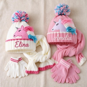 May include: Two knitted winter sets featuring beanies with pom-poms and unicorn patches, scarves, and gloves. One set is white with the name Elina, and the other is pink with the name Amelia. Both beanies feature colorful yarn pom-poms.