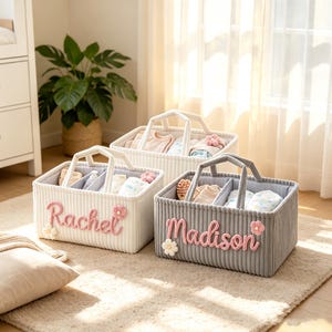 Peut inclure: Trois paniers de rangement rectangulaires en tissu côtelé avec deux poignées et des séparateurs internes. Deux paniers sont blancs et un est gris. Chacun porte un prénom personnalisé en laine rose, Rachel ou Madison, avec des fleurs au crochet.
