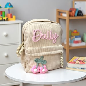 May include: Beige corduroy backpack with the name "Dally" in pink script. The backpack features a front pocket with a cherry blossom design. The bag is sitting on a white table in a child's room.