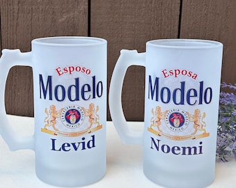 Pode incluir: Duas canecas de cerveja de vidro fosco com alças. As canecas apresentam o logotipo Modelo, a palavra "Esposo" acima da palavra "Modelo" em uma caneca e o nome "Levid" abaixo. A outra caneca tem a palavra "Esposa" acima de "Modelo" e o nome "Noemi" abaixo.