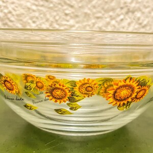 May include: Clear glass bowl with a lid, decorated with yellow sunflowers and green leaves. The bowl has the words "fleur du" and "fleur de" printed on it. The bowl is likely used for serving or storage.