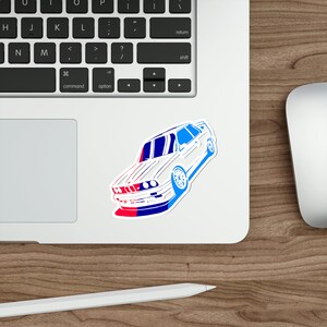 Può includere: Un adesivo per auto bianco con contorno blu e rosso. L'auto è una BMW M3 con carrozzeria bianca e una striscia blu e rossa sul paraurti anteriore.