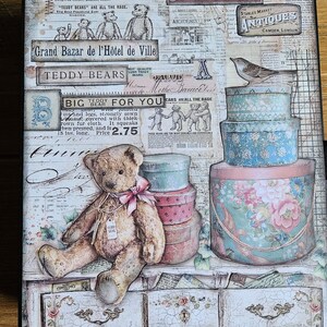 May include: A rectangular decorative board featuring a collage of vintage-style newspaper clippings, text including "Brocante" and "Teddy Bears," a brown teddy bear with a pink bow, stacked floral patterned boxes, and a small bird perched on top.