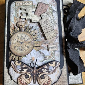 May include: A vintage-style scrapbook with a butterfly, pocket watch, and decorative paper elements on the cover. The text includes "No. 165036" and a quote. A black ribbon is tied on the side. The scrapbook has a nostalgic feel.