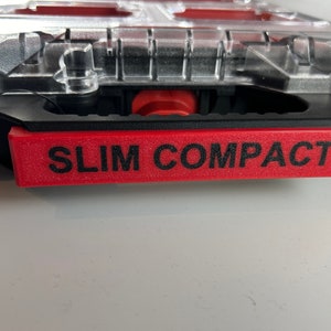 Könnte beinhalten: Ein transparentes Kunststoff-Aufbewahrungskoffer mit einem roten und schwarzen Griff. Der Koffer hat ein rotes Etikett mit der Aufschrift "SLIM COMPACT".