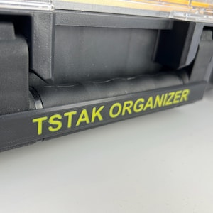 Może przedstawiać: Zbliżenie na czarny organizer na narzędzia z żółtą etykietą z napisem "Tstak Organizer".