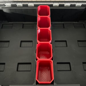 Könnte beinhalten: Fünf rote Plastikbehälter, die vertikal in einer schwarzen Werkzeugkiste gestapelt sind. Die Behälter haben eine quadratische Form mit abgerundeten Ecken.