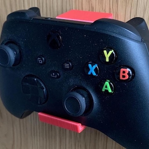 May include: A black Xbox controller with red, yellow, green, and blue buttons is mounted on a red wall mount. The controller is attached to the mount with a small red plastic piece.