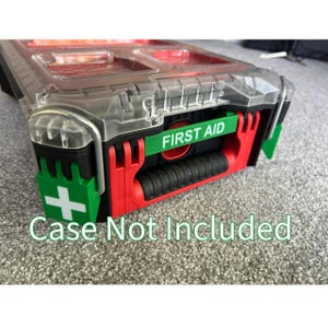 Può includere: Una cassetta degli attrezzi in plastica nera e rossa con un coperchio trasparente. La cassetta degli attrezzi ha un'etichetta verde che dice "FIRST AID" e un adesivo verde con un segno più bianco. La custodia non è inclusa.