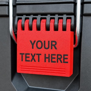 Può includere: Un'etichetta rettangolare in plastica rossa con un meccanismo a cerniera superiore fissata su una superficie nera. L'etichetta presenta il testo stampato in nero YOUR TEXT HERE al centro, con due linee orizzontali in rilievo sul bordo inferiore.