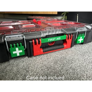 Puede incluir: Una caja de almacenamiento de plástico negro con detalles rojos y verdes. La caja tiene varios compartimentos y un asa. La caja tiene una etiqueta que dice "FIRST AID" y un símbolo de cruz verde en el lateral.