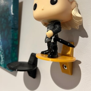 May include: A yellow wall mount shelf with a black and gray figurine standing on it. The shelf is attached to a white wall with a screw. The figurine is a pop culture character with a sword.