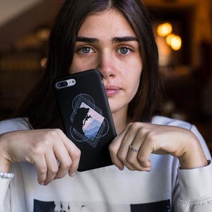 May include: Black phone case featuring a geometric design with a mountain silhouette in shades of blue and pink. The case has a sleek, matte finish and a camera cutout. The design is centered on the back of the case.