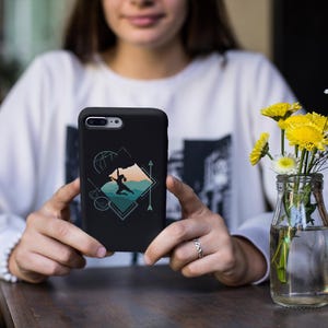 May include: Black phone case featuring a geometric design with a silhouette of a person, mountains, and a basketball. The case is held in front of a vase of yellow flowers and white baby's breath.