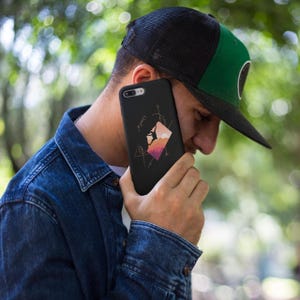 May include: Black phone case featuring a mountain climbing design with geometric shapes and a climber silhouette. The case has a colorful sunset-like background. The phone case is held by a person wearing a denim jacket and a black and green trucker hat.