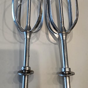 May include: A pair of silver-colored metal hand mixer beaters. Each beater features a central rod with four curved wire loops forming an oval cage at the top, a circular collar, and a notched base for insertion into a kitchen appliance.