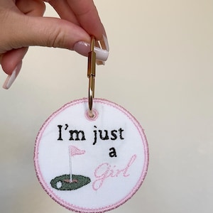 May include: Embroidered keychain with the text "I'm just a Girl" and a golf-themed design. The round patch is white with pink stitching and a gold-coloured clasp. The design includes a golf flag and ball.