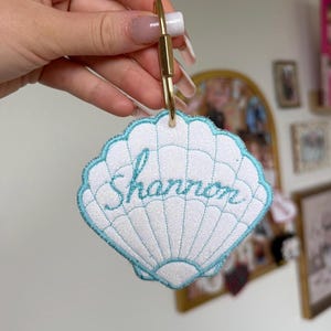 May include: White seashell-shaped keychain with the name "Shannon" embroidered in blue script. The keychain has a gold-colored clasp and a light blue scalloped border. The seashell design is detailed with stitched lines.