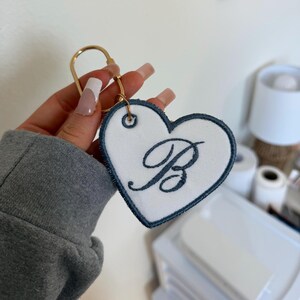 Puede incluir: Llavero en forma de corazón con centro de tela blanca y borde bordado en azul oscuro. La letra "B" está bordada en una fuente cursiva. El llavero tiene un anillo de metal dorado.