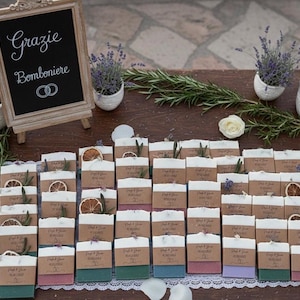 Puede incluir: Exhibición de recuerdos de boda, incluyendo pequeños jabones con rodajas de naranja seca y ramitas de romero. Un letrero de pizarra dice "Grazie Bomboniere" con anillos de boda. Plantas de lavanda y olivo decoran la mesa.