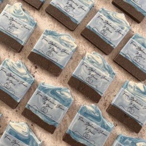 May include: Artisan soaps with a marbled blue and white design, wrapped in paper with the text "From Stephanie's Shower To Yours" and two wedding rings. The soaps have a dark brown base and are arranged on a light-colored surface.