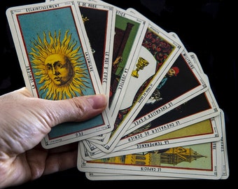 May include: A fan of tarot cards featuring French text, including La Lumière, Le Roi d'Épée, La Justice, Le Trois de Coupe, Le Chevalier d'Épée, Le Chevalier de Denier, La Tempérance, and Le Capucin, with a prominent sun illustration on the first card.