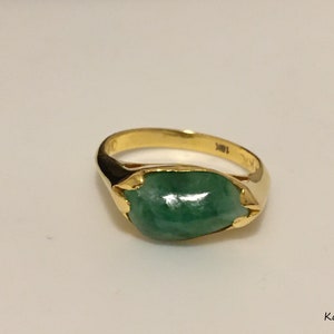 May include: A gold ring featuring a smooth, polished, oval-shaped green jade stone set in a yellow gold band. The interior of the band is clearly stamped with the text 18K. The text KaysJade is visible in the bottom right corner of the image.