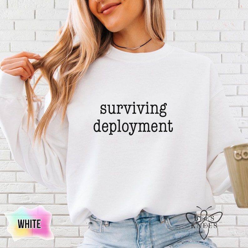 May include: White sweatshirt with the text "surviving deployment" in a black font. The sweatshirt is a crew neck style. The word "WHITE" is in a pink and blue gradient box in the lower left corner.