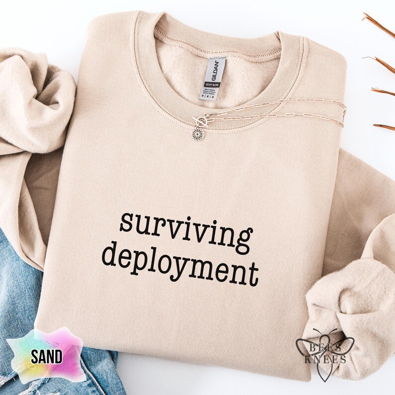 May include: A sand-colored sweatshirt with the words "surviving deployment" printed in black. A delicate silver necklace with a small pendant rests on the sweatshirt. The sweatshirt is folded, with a portion of denim jeans visible.