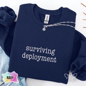 May include: Navy blue sweatshirt with the text "surviving deployment" in white. A silver necklace with a sunburst pendant is draped across the sweatshirt. The sweatshirt is folded on a white surface.