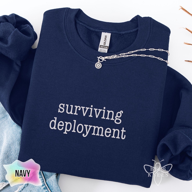 May include: Navy blue sweatshirt with the text "surviving deployment" in white. A silver necklace with a sunburst pendant is draped across the sweatshirt. The sweatshirt is folded on a white surface.